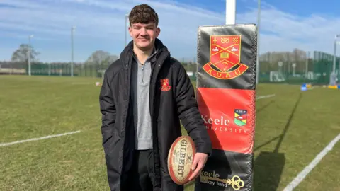 Keele University Spencer has brown hair and is wearing a grey top and black coat. He is holding a white and red rugby ball. He is standing on a green rugby pitch with a rugby post behind him, that has a padded material over it with the Keele University logo on a red background