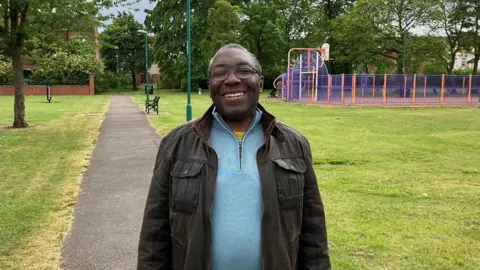 Dominic Nelson-Ashley standing in Ayresome Gardens. He is smiling as he looks into the camera. He has short greying hair and is wearing black glasses, a brown jacket and blue top. Behind him are grassed areas and a basketball court.