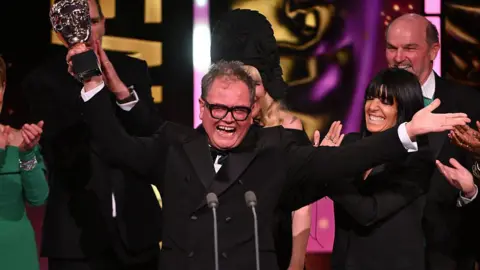 Getty Images Comedian Alan Carr in a dark suit with his trademark large glasses holds a Bafta award in one hand with his arms opened wide in celebration. Claudia Winkleman is beside him.