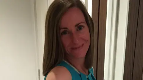 A woman in a blue dress is smiling, posing for the camera. She has brown hair and is stood next to a mirror.