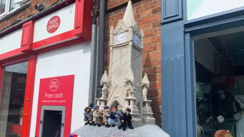 Knitted postbox topper with knitted figures doing a conga around Leicester's clock tower.