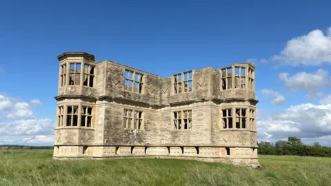National Trust Images/Kate Scott Lyveden Lodge, a roofless late 16th Century building built out of cream-coloured stone, showing two sides of its cross shape. It is surrounded by grass and above is a blue sky. 