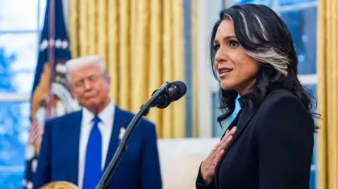 Gabbard with her hand over her heart speaking in front of a microphone in the Oval Office, while Trump can be seen out of focus in the background