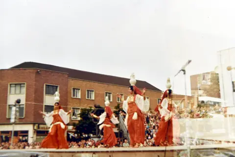 Walter Crinnion Woman in red dresses dancing on stage with white vases on their heads. Crowds of people look on. A three-storey brick building with shops on the ground floor is in background.