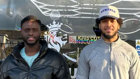 Two men stood outside a barber shop, one is wearing a black leather jacket, the other is wearing a grey 3/4 zip sweater and a white baseball hat. Both men have short brown curly hair and are smiling at the camera