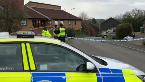 BBC Three police officers are standing behind a police car next to tape across the road