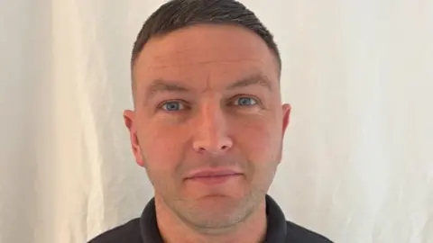 Michael Harkin Michael Harkin has short brown hair. He is wearing a black polo shirt and is standing in front of a white background