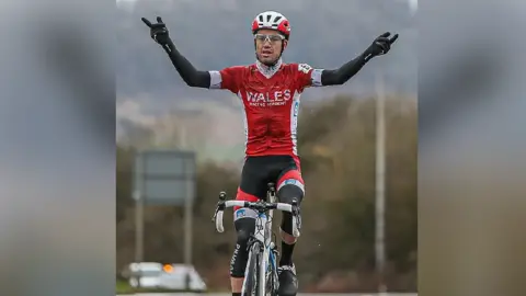 Huw Fairclough Peter Kibble on his bike with his hands in the air. He is wearing a red cycling outfit with Wales written in white letters on the front. 