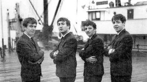Peter Kaye Photography Image shows George Harrison, John Lennon, Ringo Starr and Paul McCartney in 1962, standing on the landing stage at Liverpool, with the distinctive mast of the Salvor behind them. It has three masts that point outwards on an angle. 