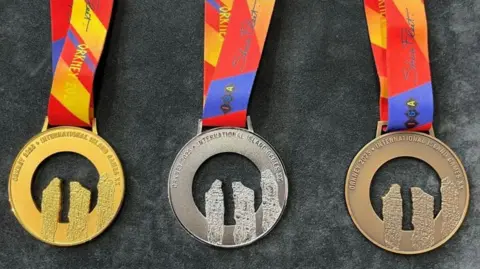 Gold, silver and bronze medals, which are circular shaped and have the shape of three standing stones cut out of them. They are attached to colourful ribbons.