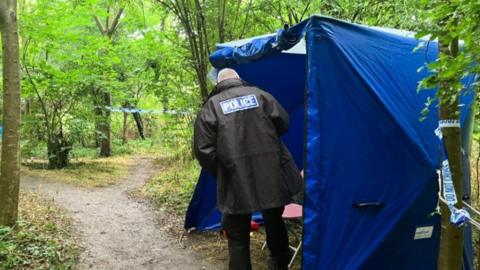 Trees line a narrow path that forks off in two different directions in the background. In the foreground there is a blue police tent with the back view of police officer in black coat, standing at entrance to the tent