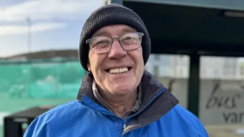 Joe Edge is smiling, wearing glasses and wearing a black woolly hat. He is wearing a bright blue waterproof jacket and standing in front of a bus shelter.
