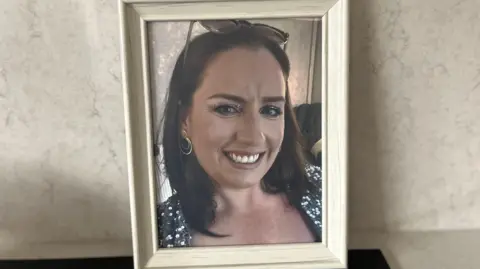 A white photo frame contains the photo of a woman with long brown hair, who wears a black and white floral top. She smiles whilst wearing multiple golden hooped earrings. The frame is propped up against a cream wall. 
