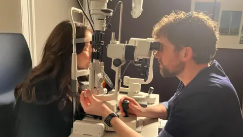 Edinburgh Innovations Optometrist Ian Cameron giving a woman an eye examination through a piece of specialised equipment. He is looking into the machinery on one side as she rests her chin on the either side as he looks into her eye. One of her eyes is lit up.