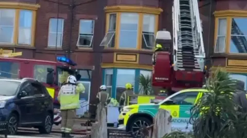 Shakar Bandyan Fire crews outside a building which has bay windows and is made of red brick. There are two fire engines visible and an emergency car. One fire engine has its ladder extended towards the buildings.