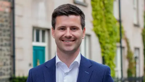 PA Media Buckley is smiling at the camera. He has short brown hair and is wearing a navy pinstripe suit jacket and a white shirt without a tie. 