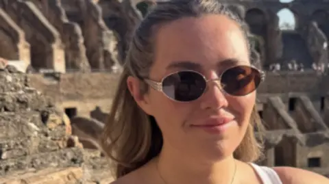 Ella Nickson is wearing sunglasses and her hair is half-tied up. She is standing inside the Colosseum in Rome