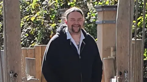 A man in a shirt and black hooded top.  He has long hair and is smiling. He is standing on a wooden bridge