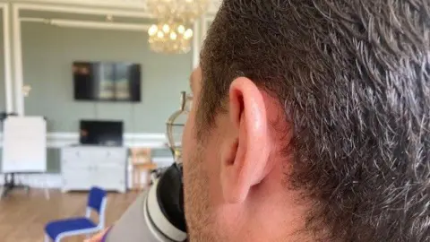 Photo of a man, only showing the back and side of his head, sipping drink from a cup. He has dark hair and wears glasses.