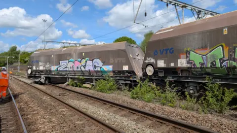 A wagon has come off a track and sits on another neighbouring line. Two wagons can be seen in the image, both what looks like a dirty brown or metal colour with graffiti on. Some rail workers stand nearby. 