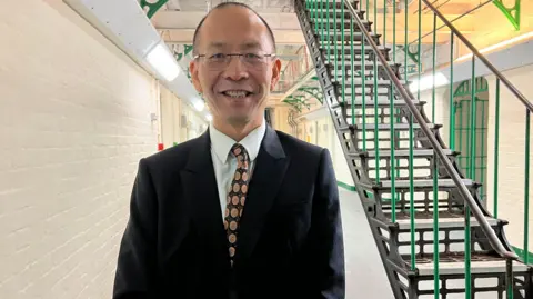 A Chinese man wearing a black suit, white shirt and yellow and black patterned tie smiles towards the camera. Behind him are a dark green iron staircasr and cream painted brick walls