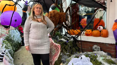 BBC Claire Cox, a woman with long blonde hair in a grey dress, stands outside her house covered in Halloween decorations