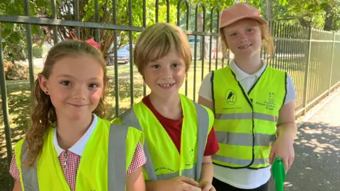 Three children wearing yellow high-vis vests are standing outside their school gates smiling at the camera.