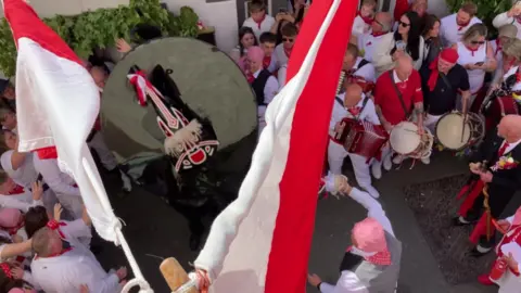A crowd of people cheer and sing as a large black Obby Oss with red ribbons dances through the streets of Padstow, surrounded with flags 