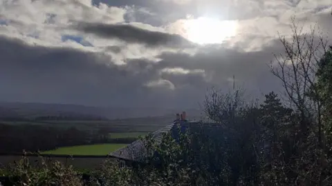 Weather Watchers/Pauls Eye A dark, cloudy sky with the sunshine peeping through dramatically over a landscape of fields with a house slate roof in the foreground.
