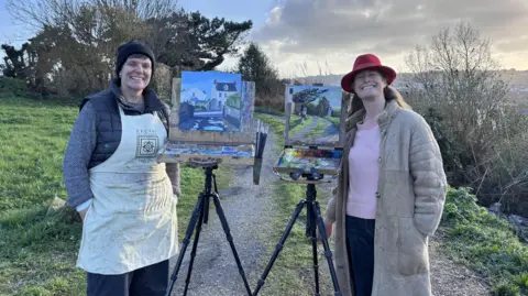 Rosanne Guille (left) and her friend Susanna MacInnes (right) next to their landscape paintings which are on easels. They are both standing on a path with grass and trees on either side. Both women are looking straight at the camera and both are smiling.