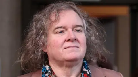 A middle aged woman with long curly grey hair wearing a colourful scarf
