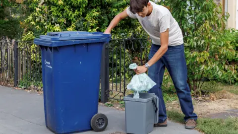 David Johnson A young man is putting a plastic bag of waste into a small grey bin. There is a larger blue bin on the pavement next to it. The man is wearing a cream T-shirt and blue jeans.