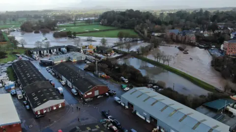 The River Otter overtopping in Ottery St Mary and flowing into surrounding fields, close to homes and businesses.