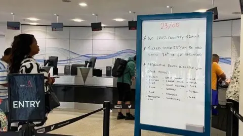 A hand-written whiteboard details the cancellations to trains, in the customer ticket office. On the right there is a sign that says Entry, behind which passengers are waiting to go to the counter.