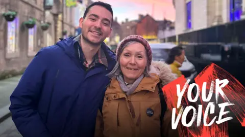 Deborah Crowley Deborah and her son Stuart smile while wearing coats - Stuart is wearing a royal blue one and hers is a burnt orange. They are standing in the street, with a pink sunset in the sky behind them. In the bottom right corner of the image is a white and red 'Your Voice' logo.