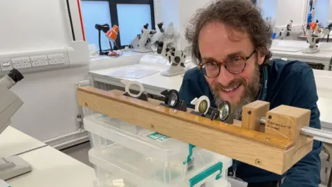 A man in a blue shirt and glasses smiles. He is looking at an array of optician equipment on a brown board