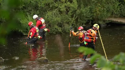 Search and rescue team of four in the River Tyne in Wylam. Two are wearing red helmets, one has a yellow helmet and the fourth is in white. They are wearing red and yellow waterproof clothing and red lifesaver jackets. Each has a pole with which they appear to prodding into the water, which is a murky green. Beyond them and in the foreground are trees, shrubs and other vegetation.