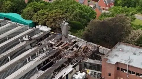 A picture taken with a drone, showing part of the fire-damaged factory, with homes behind it, divided by trees.