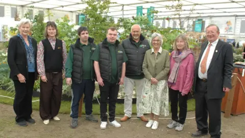 A group photo of two members of Grow standing alongside six members of the Cornish-based Burncoose Nurseries. In the background is the garden.