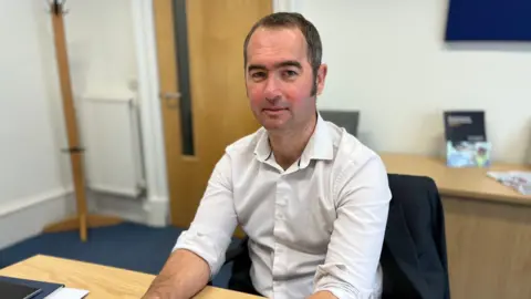 BBC News Scott McIver, with short, dark hair, wearing a white shirt with rolled up sleeves, sitting at a desk in an office 