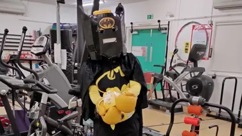 Julianne Moroz A woman who is dressed up in a Batman costume, with a black cape, a black bat head. They are standing in a gym and are wearing yellow boxing gloves which have a picture of Pudsey Bear attached to them. Weightlifting machines are in the background. 