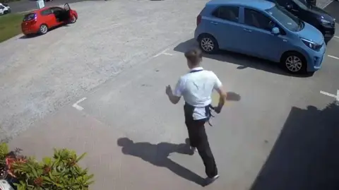 Young man wearing a white shirt and apron, running through a car park towards a red car