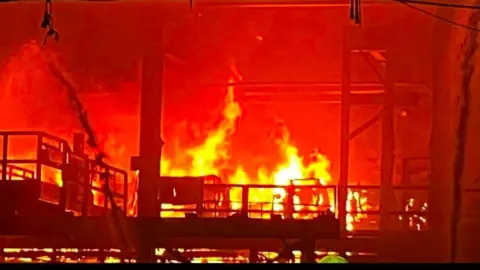 Close up image showing a large fire burning tn the steelworks with large yellow flames. 