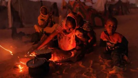 Getty Images A woman in a refugee camp cooks at an open fire with the light from the flames illuminating her face. A child sits to her left. 