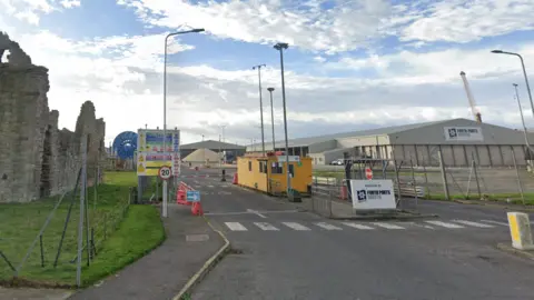 Google Entrance to Rosyth Port showing security gates, signage, and industrial buildings, with the ruins of an old stone structure on the left and a partly cloudy sky overhead.