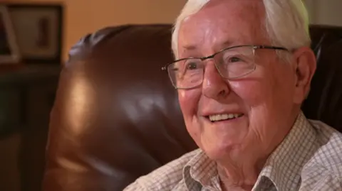 Stephen Harrington is an older man, aged 83. He has white hair and wears thin-rimmed clear-lensed glasses. He is sitting on a brown leather chair and wears a white and brown checked shirt. He is smiling.