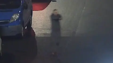 COPFS A grainy CCTV image of Hunter with his arms stretched out in front of him holding a gun. He is standing upright on a road in grey clothing. A blue van is passing behind him.