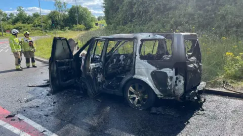 A car destroyed by fire, with two firefighters standing by it