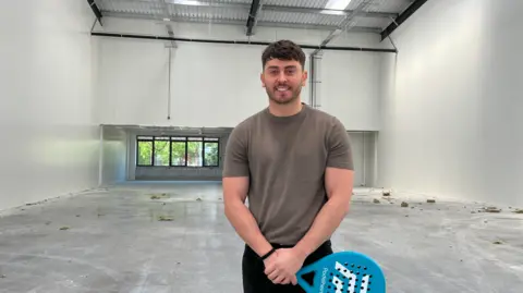 BBC A man in a brown T-shirt with dark hair is smiling at the camera and holding a racquet. He is standing inside in a big empty space with white walls and windows in the background beneath a large wall.