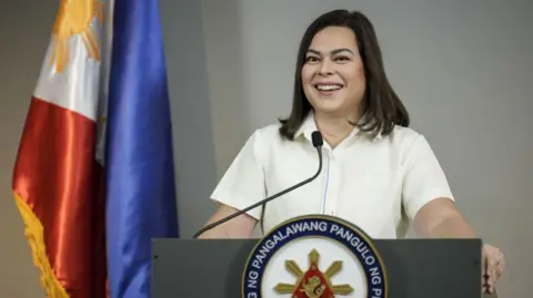 Getty Images Sara Duterte stands behind a podium and addreses the media in a white shirt, smiling. Behind her is the Philippines flag.